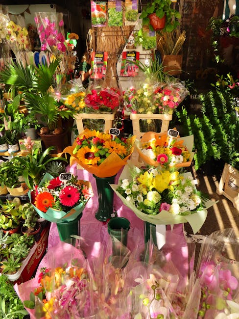 A vibrant display of fresh flower bouquets arranged on a pink cloth-covered table within a flower shop. The bouquets include bright yellow sunflowers, pink gerbera daisies, red roses, and white chrysa