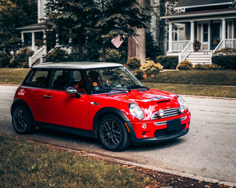 A vibrant red Mini Cooper with a black roof and black alloy wheels parked on a residential street in front of a suburban house with white exterior walls and a porch. The car's sleek and shiny exterior