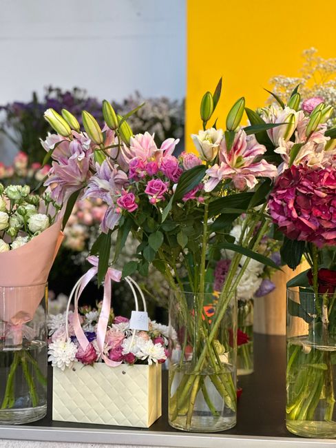 A display of fresh floral arrangements featuring pink lilies with elongated buds and pale pink petals, complemented by clusters of small pink roses and dark green leaves. The flowers are arranged in c