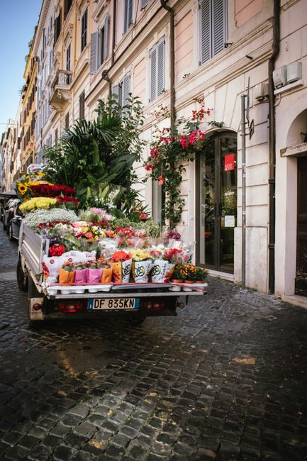 A delivery truck parked on a cobblestone street fronting a classic European building with cream-colored walls and green window shutters. The truck's open flatbed is filled with a vibrant variety of fr
